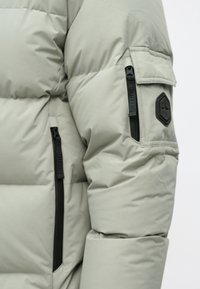 Vaaleanharmaa untuvapuku, jossa on tikattu muotoilu, teksturoitu kangas, vetoketjullinen tasku ja kuusikulmainen logolappu hihassa.