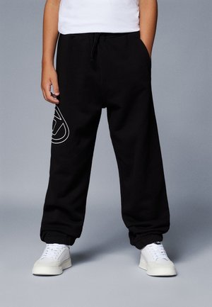 Enfant portant un pantalon de survêtement noir avec un logo blanc sur la jambe gauche, un t-shirt blanc et des baskets blanches, debout devant un fond gris.