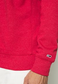 Sudadera roja con un patrón de tejido texturizado, puños acanalados y un parche de logo en la manga. Cuenta con un cuello redondo y un dobladillo elástico.