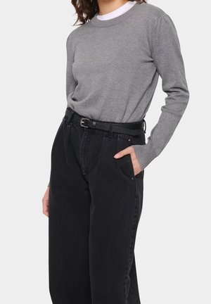 Woman wearing a gray long-sleeve sweater tucked into high-waisted black jeans with a black belt, one hand in pocket.