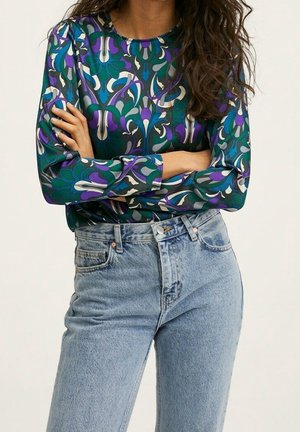 Kobieta z długimi kręconymi włosami, ubrana w kolorową, kwiecistą bluzkę z długim rękawem oraz jasnoniebieskie jeansy z wysokim stanem, stojąca z założonymi ramionami.