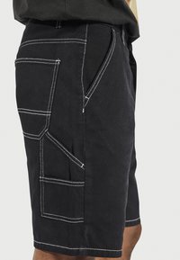 Schwarze Shorts mit mehreren weißen, aufgenähten Taschen, getragen von einer Person, die Seiten- und Rückansicht sowie kontrastierende Nähte zeigt.