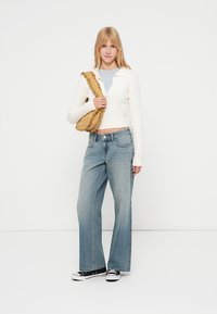 Pulover scurt alb cu model tricotat, bluză albastru deschis, blugi largi decolorati și adidași negri. Geantă galbenă cu volane purtată pe umăr.