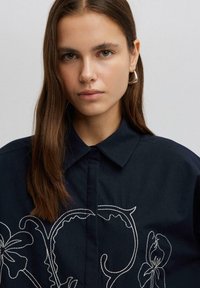 Camicia blu navy con colletto, caratterizzata da ricami floreali bianchi sulla parte anteriore, realizzata in un tessuto liscio. Il design è dettagliato e audace.