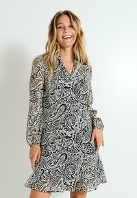 Vestido de patrón paisley en blanco y negro con mangas largas abullonadas, escote en V y un dobladillo con volantes. Material transparente con una silueta fluida.