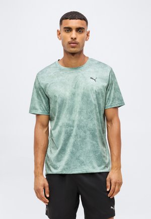 Mężczyzna ubrany w wyblakły zielony t-shirt Puma i czarne szorty Puma stoi na tle jednolitego jasnego tła.