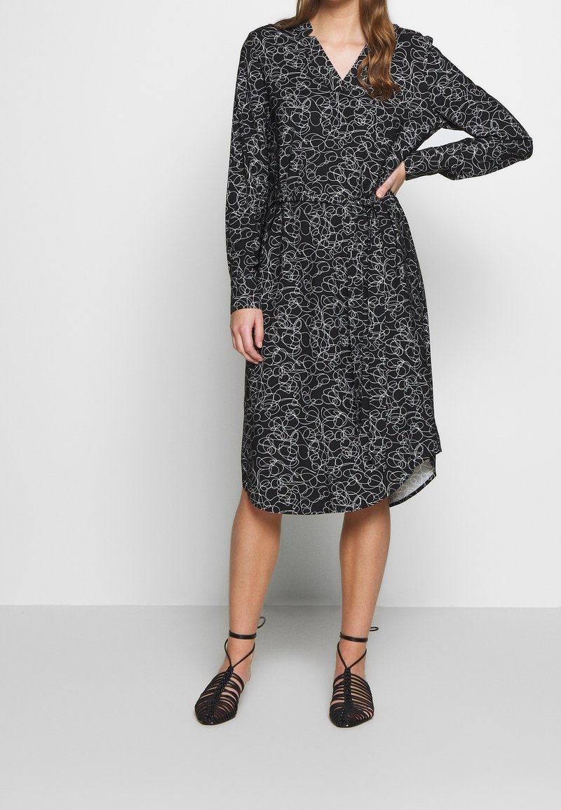 Femme portant une robe noire longueur genou avec des motifs abstraits en spirale blancs et des chaussures plates noires à brides, debout avec une main sur la hanche.