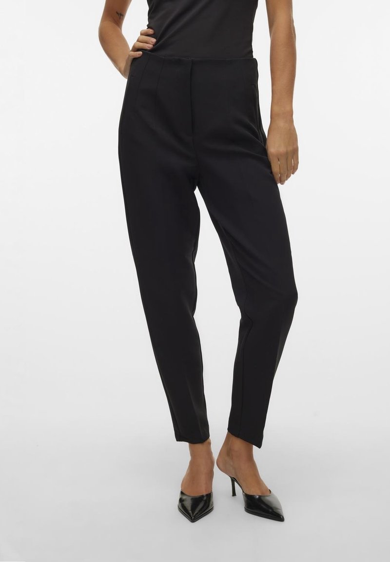 Femme portant un pantalon noir slim taille haute assorti à des escarpins noirs à bouts pointus, debout avec une main sur la hanche devant un fond uni.