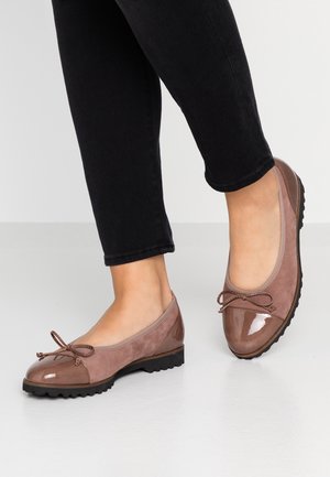 Fødder iført brune ballet flats i ruskind og laklæder med sløjfedetalje, kombineret med sorte ankellange bukser på et hvidt gulv.