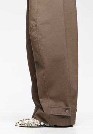 Brown wide-leg trousers partially covering pointed-toe snakeskin-patterned shoes against a plain background.