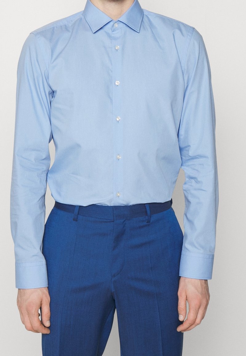 Homme portant une chemise bleue claire boutonnée, rentrée dans un pantalon formel bleu foncé avec passants de ceinture, debout devant un fond neutre.
