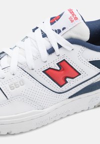 Fehér sportcipő piros és kék részletekkel, perforált részletekkel, vastag fehér fűzőkkel, valamint a "550" és a "New Balance" felirattal.
