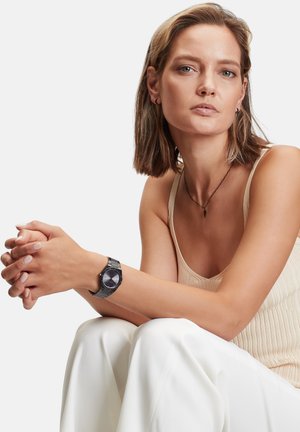 Herrenarmbanduhr mit schwarzem runden Zifferblatt, silbernem Metallarmband und minimalistischen Design. Modell trägt ein beiges Oberteil, sitzt mit gefalteten Händen.