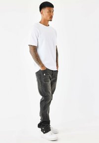 Witte crew-neck t-shirt, zwarte distressed jeans en witte sneakers. Kenmerken zijn onder andere tatoeages op de armen en een ontspannen pasvorm van de kleding.
