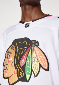 Valge hokipusa, millel on värvikas tikand Native American profiiliga, millel on sulgede aktsendid ja NHL-i märk kaeluses.