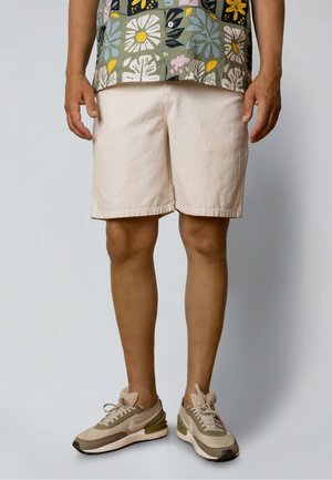 Man die beige shorts, beige en olijfgroene sneakers en een bloemenpatroon overhemd draagt, staande tegen een effen achtergrond.