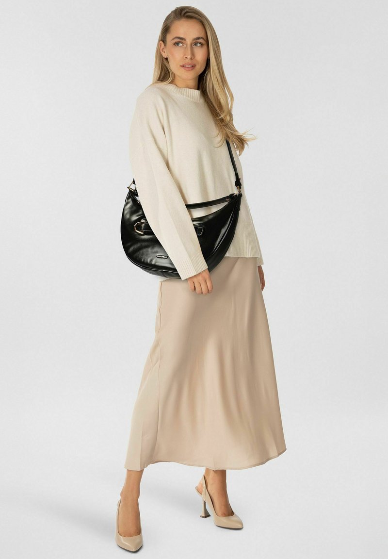 Borsa a mano in pelle nera con forma curva, tracolla laterale e dettagli in oro, abbinata a un maglione in maglia color crema e una gonna in raso beige.