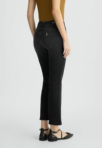 Jean en denim noir avec taille haute et jambe droite, comprenant deux poches arrière et une fermeture éclair. Porté avec des chaussures plates noires.