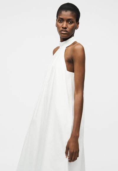 Femme aux cheveux courts portant une robe blanche sans manches à col licou, debout devant un fond clair uni, regardant vers la caméra.