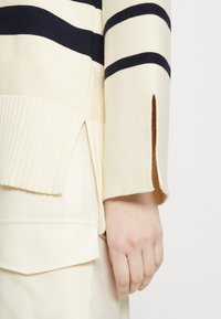 Strickpullover aus cremefarbenem Stoff mit marineblauen horizontalen Streifen, seitlichen Schlitzen und geripptem Saum. Die Hand ruht auf hellfarbigen Hosen.