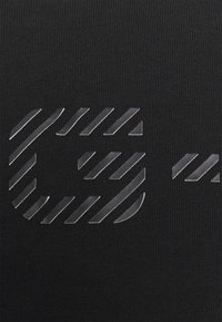Černá látka s texturovaným povrchem obsahuje logo vyrobené z lesklého, průsvitného materiálu s diagonálními pruhovanými akcenty.