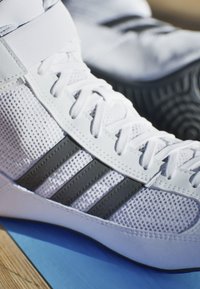White athletic shoe with mesh upper, black three-stripe logo, and rubber sole. Laces are white; overall design is streamlined and modern.