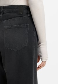 Czarne spodnie dżinsowe z teksturowanym wykończeniem, z pojedynczą tylną kieszenią oraz czarną metką z logo BOSS na pasie. Wyraźne szwy i przeszycia.