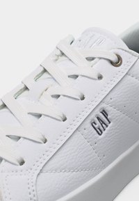 Baskets en cuir blanc avec lacets blancs, œillets argentés et logo "GAP" embossé sur le côté près de la semelle.
