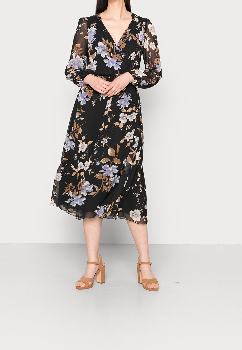 Robe noire à fleurs avec manches longues, décolleté en V et ourlet mi-long. Présente de grandes fleurs violet clair et brunes. Associée à des sandales à talons hauts couleur beige.