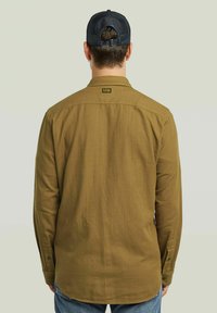 Uomo che indossa una camicia a maniche lunghe verde oliva e jeans blu, girato di spalle, con un cappellino in rete nera su uno sfondo neutro chiaro.
