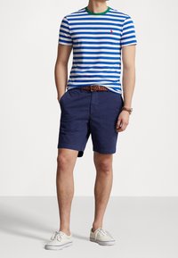 Páskované tričko s modro-bílými horizontálními pruhy, zeleným kulatým výstřihem a červeným logem. Spárované s námořnickými šortkami a bílými plátěnými botami.