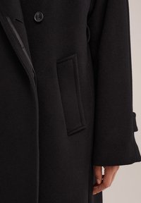 Schwarzer Wollmantel mit Reverskragen, einem einzigen Knopfverschluss und seitlichen Taschen, der eine angenehme Textur und ein strukturiertes Design aufweist.