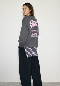 Szürke, túlméretezett pulóver rózsaszín grafikus felirattal a háton, csíkos felső fölött, széles fekete nadrággal párosítva.