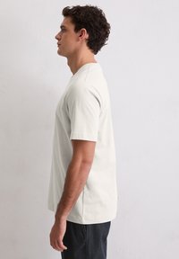 Hellgraues T-Shirt mit kurzen Ärmeln aus weicher Baumwolle, versehen mit einem Rundhalsausschnitt und einer lockeren Passform. Kombiniert mit dunklen Jeans.