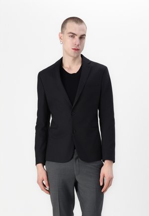 Black tailored blazer with two buttons and side pockets, paired with a black t-shirt and gray trousers. Smooth fabric and fitted design.