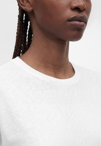 Gros plan d'une personne aux cheveux tressés portant un t-shirt blanc uni à col rond, montrant le cou et la ligne de la mâchoire sur un fond uni.