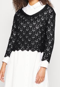 Svart stickad kort tröja med öppen spetsdesign och vågig kant, bärs över en vit skjorta med knappar och långa ärmar.