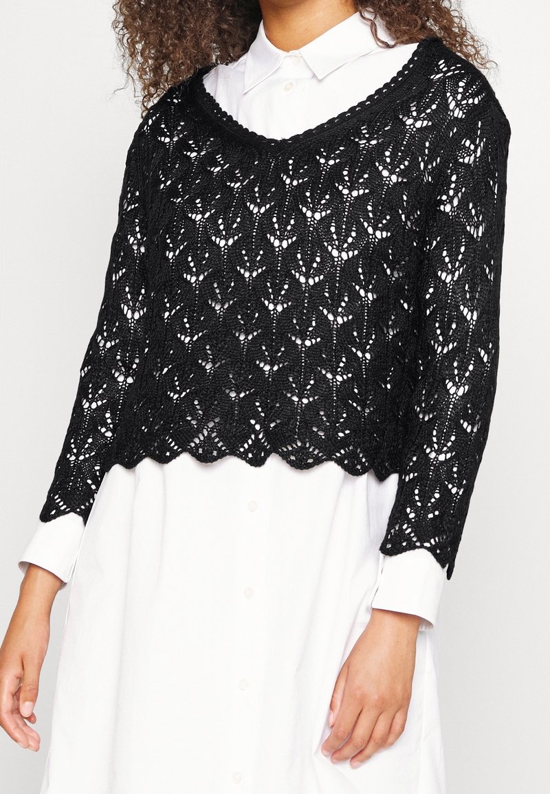 Svart stickad kort tröja med öppen spetsdesign och vågig kant, bärs över en vit skjorta med knappar och långa ärmar.