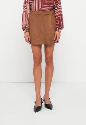 Kobieta w brązowej zamszowej mini spódnicy, bluzce z długim rękawem o geometrycznym wzorze oraz czarnych czółenkach typu slingback z szpiczastym noskiem, stojąca na białym tle.