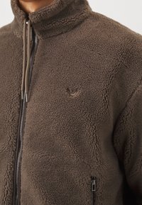 Brązowa kurtka z polaru z wysokim kołnierzem, suwakiem z przodu, troczkami i małym logo na piersi po lewej stronie.