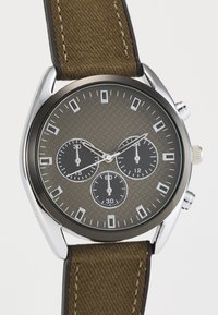 Montre argentée avec un cadran gris texturé, trois sous-cadrans et un bracelet en tissu brun. Comprend des index horaires proéminents et une couronne.