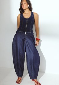 Denim overall s srajco brez rokavov, z zadrgo spredaj in širokimi, ohlapnimi hlačami. Temno modra tkanina, kontrastni šivi in drzni dodatki.