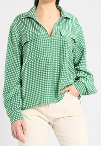 Chemise à carreaux verts à manches longues, col et deux poches avant. Tissu léger avec une coupe décontractée et un ourlet qui descend légèrement.