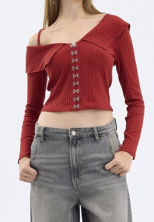Kobieta ubrana w prążkowany, czerwoną bluzkę z odkrytymi ramionami i zapięciami na haftki oraz szare, wysokowalne dżinsy, z rękami w kieszeniach.