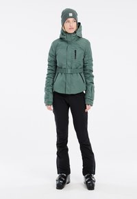 Groene geïsoleerde jas met een capuchon, elastische riem en ritszakken, gecombineerd met zwarte broeken en zwarte sneeuwlaarzen. Gestructureerde stof.