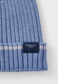 Blå stickad mössa med ribbat mönster och vita ränder. Har en marinblå etikett med "HACKETT LONDON" logotyp. Mjuk garnmaterial.