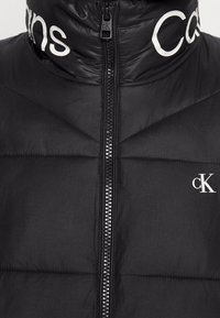 Chaqueta acolchada negra con cuello alto, con logotipo blanco, cremallera frontal y detalles acolchados en toda la tela.