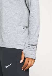 Grijs sportief longsleeve shirt met een gestructureerde afwerking, voorzien van een zijzak en duimopeningen. Gecombineerd met zwarte broek en een Nike-accent.