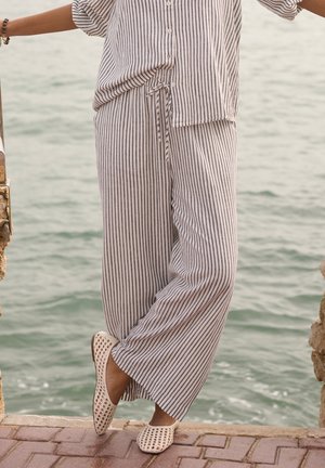 Persona che indossa pantaloni larghi a righe e camicia abbottonata coordinata, ferma vicino all'acqua su un pavimento in mattoni, indossa scarpe beige intrecciate senza lacci.