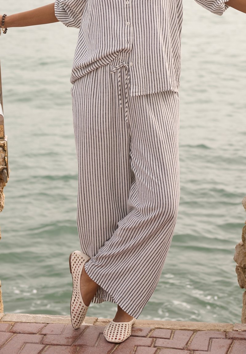 Personne portant un pantalon ample à rayures et une chemise boutonnée assortie, debout près de l'eau sur un pavé en briques, portant des chaussures beiges tissées sans lacets.